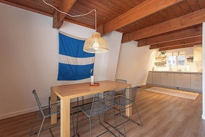 Uma sala de jantar com mesa de madeira e cadeiras de arame, luminária pendente e cozinha ao fundo.