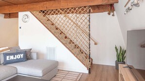 A room with a wooden staircase, a sofa with a cushion labeled 'ENJOY', a potted plant, and a wall-mounted clock. - Ocean Pines 2BR 1BA Cottage with Fire Pit (Ocean Pines)