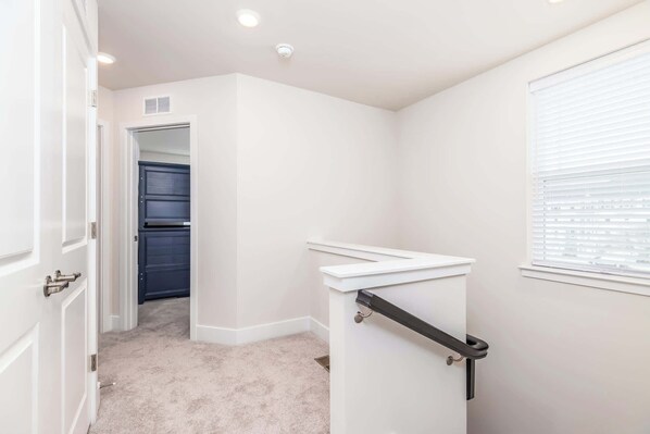 A hallway with a white door, a dark closet, a white wall, a window with blinds, and a white staircase with a black handrail. - New Townhome Steps from the Beach (Ocean City)