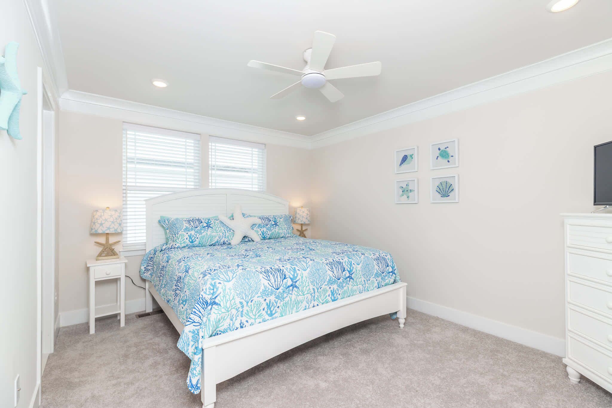 A bedroom with a bed, a ceiling fan, a nightstand with a lamp, and wall art.