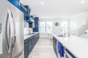 A modern kitchen with a stainless steel refrigerator, dark blue cabinets, a white countertop, and a window with blinds. - New Townhome Steps from the Beach (Ocean City)