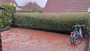En asfalteret indkørsel fører til et hus med rødt tag, en hæk og en cykel parkeret ved siden af.
