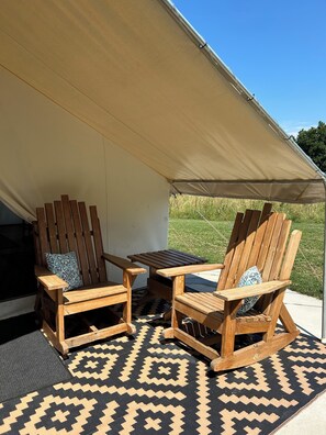 Un espace extérieur aménagé avec une tente, comprenant deux chaises en bois et une table, un tapis à motifs et une vue sur un champ verdoyant.