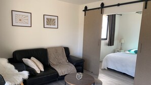 A modern living room with a black leather sofa, a round wooden table with a glass and a pearl, and a grey rug. A doorway leads to a bedroom with a bed and a nightstand.