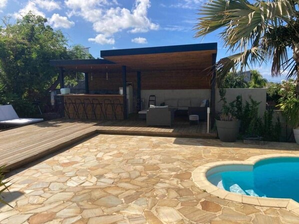 A backyard with a stone patio, a swimming pool, outdoor furniture, and a covered seating area.