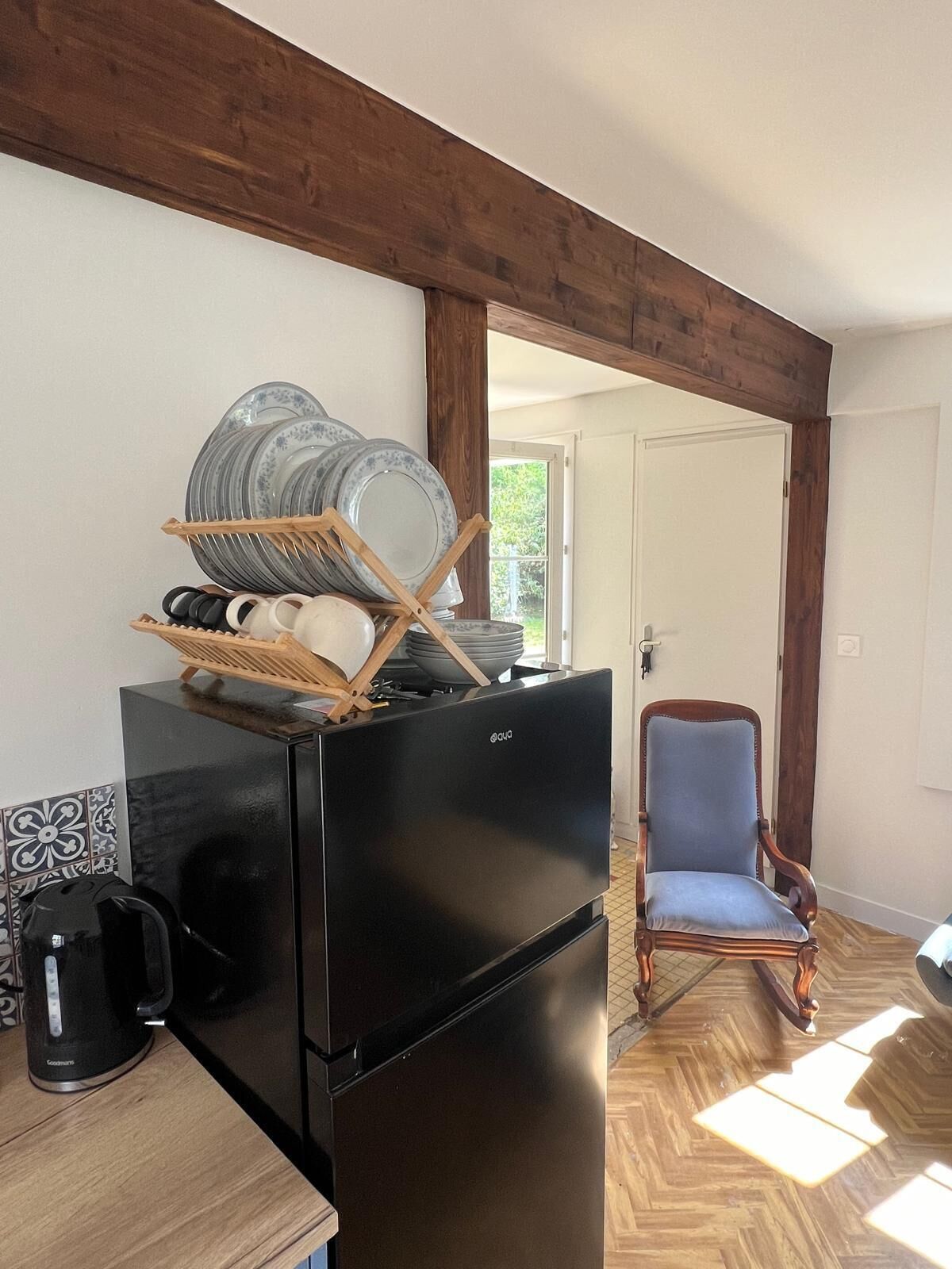 A kitchen with a black refrigerator, a coffee maker, and a dish rack with plates and bowls. There's a wooden chair and a door leading outside.