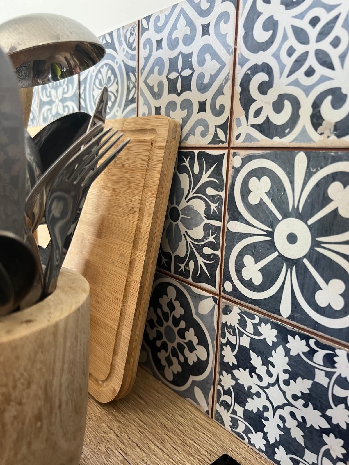 A kitchen counter with a wooden cutting board, a metal utensil holder, and a tiled backsplash with a floral pattern.