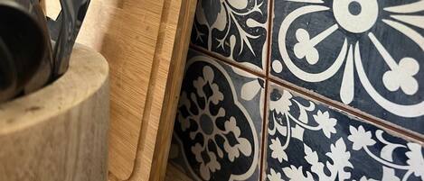 A kitchen counter with a wooden cutting board, a metal utensil holder, and a tiled backsplash with a floral pattern.