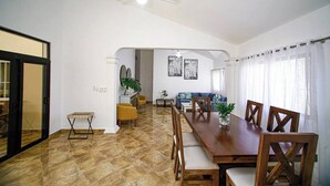 Uma sala de jantar ampla com uma longa mesa de madeira e cadeiras, um ventilador de teto e vista para uma sala de estar com um sofá e uma planta.