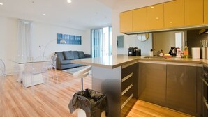 A modern kitchen with a central island, a dining area with a glass table and chairs, and a living area with a sofa and a painting on the wall. - Amyra Residences on Rowlands Place - Unit 1006 (Adelaide)
