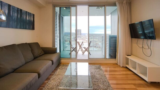 A modern living room with a sofa, a glass coffee table, and a TV mounted on the wall. - Amyra Residences on Rowlands Place - Unit 1006 (Adelaide)