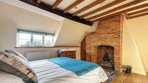 Quarto com teto inclinado, lareira de tijolos, cama com cobertor azul e janela com vista para vegetação.