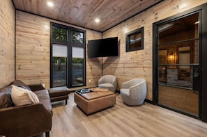 Un salon moderne avec un intérieur en bois, un téléviseur à écran plat fixé au mur, un canapé en cuir marron, deux fauteuils et une porte en verre donnant sur une cheminée en pierre.