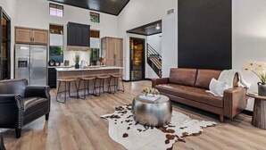 Et moderne køkken med en central ø, et spiseområde med barstole, en lædersofa og en trappe til en etage ovenpå.