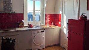 A compact kitchen with a washing machine, a red refrigerator, and a window with blinds. - Cabourg fan (Cabourg)