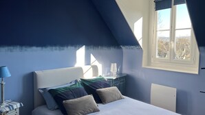 A bedroom with a sloped ceiling, a bed with multiple pillows, a nightstand with a lamp, and a window with a view of trees. - Cabourg fan (Cabourg)