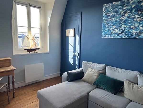A room with a grey sofa, a wooden table, a radiator, and a window with a sailboat model.