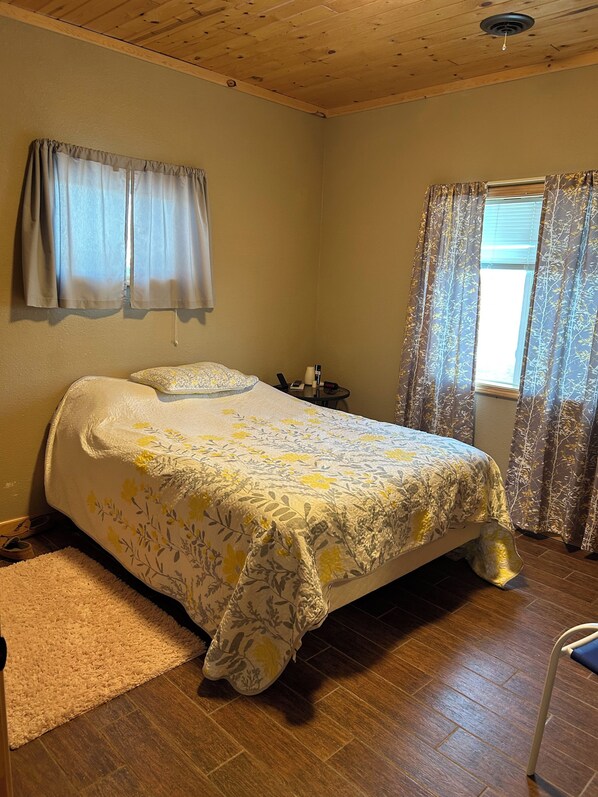Un dormitorio con cama, mesita de noche, ventana con cortinas y techo de madera.