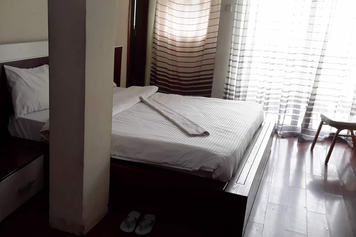 A neatly made bed with a white comforter and a folded towel, a wooden nightstand with a drawer, a pair of flip-flops on the floor, and a chair by the window with striped curtains.
