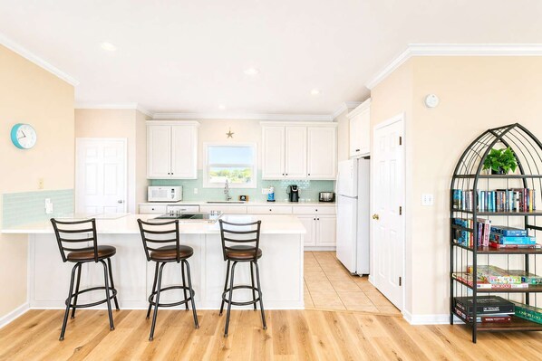 A modern kitchen with white cabinets, a central island with bar stools, a coffee machine, and a bookshelf with board games. - Barbara Ann's Happy Place (Eastpoint)