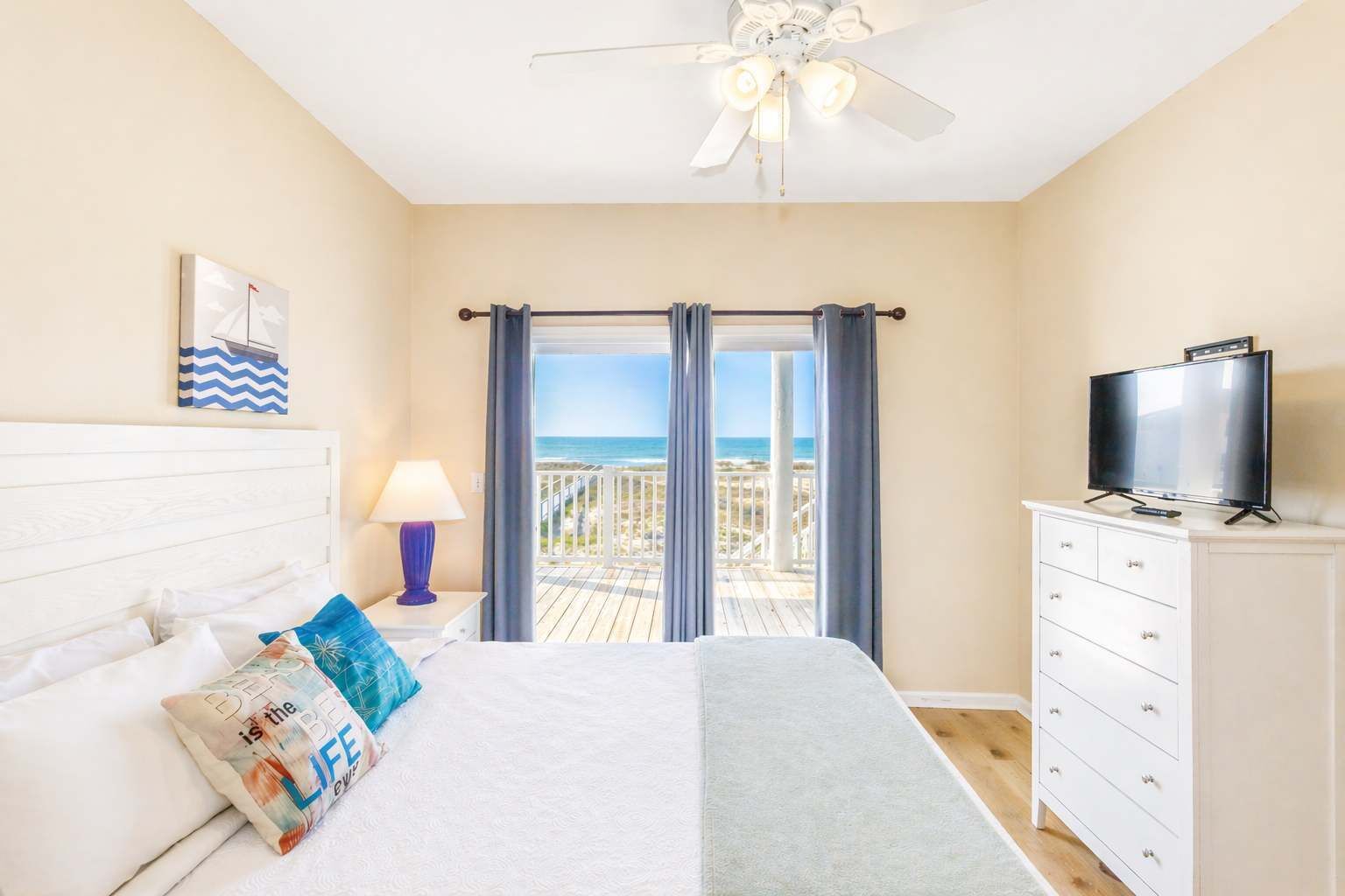 A bedroom with a bed, a TV, a ceiling fan, and a balcony with a view of the beach.