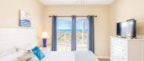 A bedroom with a bed, a TV, a ceiling fan, and a balcony with a view of the beach.