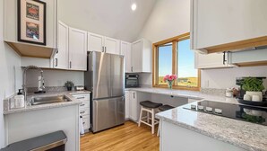 A modern kitchen with white cabinets, a stainless steel refrigerator, a built-in oven, and a window with a view of the countryside.