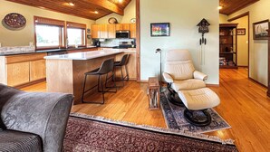 A spacious kitchen with wooden cabinets, a central island with bar stools, a large window, and a cozy seating area with a chair and a sofa.
