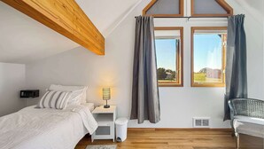A bedroom with a sloped ceiling, a bed, a nightstand with a lamp, a chair, and a window with curtains.