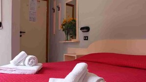 A hotel room with a red bedspread, neatly folded white towels on the bed, a small bedside table with a vase of flowers, and a mirror on the wall. - Hotel Laura Beach (Rimini)