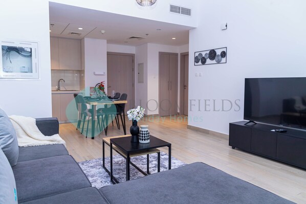 A modern living room with a sofa, a TV stand, and a dining area with a table and chairs. - Bloomfields Elegant 1BR in Waters Edge (Abu Dhabi)