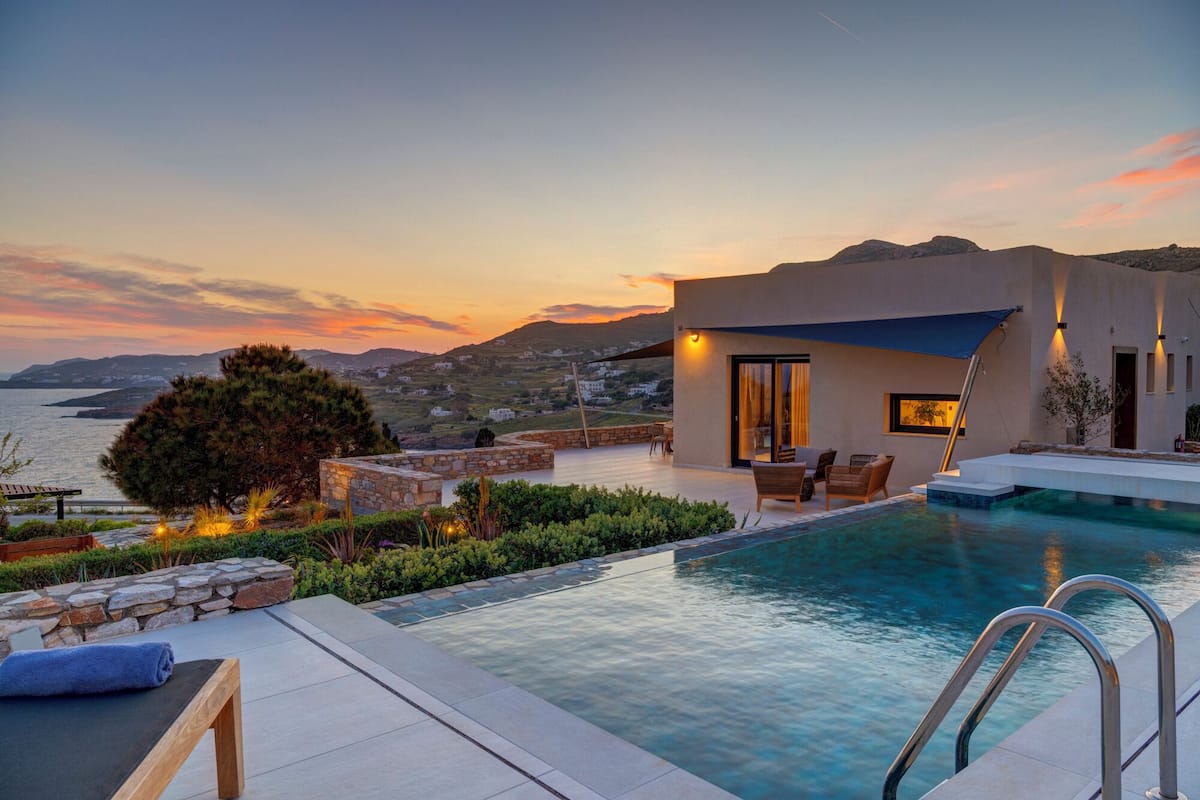A modern villa with a swimming pool, outdoor seating, and a view of the sea and hills.