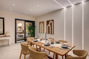 A modern dining room with a long wooden table set for six, surrounded by wicker chairs, and a glass partition separating the dining area from a bedroom.