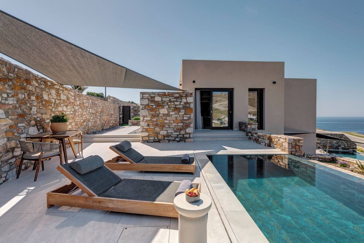 A modern outdoor pool area with lounge chairs, a stone wall, and a view of the sea.