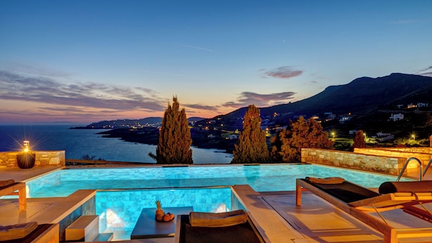 Une piscine bordée de chaises longues, un mur de pierre et une vue sur la mer et les montagnes au crépuscule.
