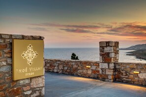 Mur de pedra amb un cartell que diu 'Pnoé Villas Syros' i una vista del mar al capvespre.