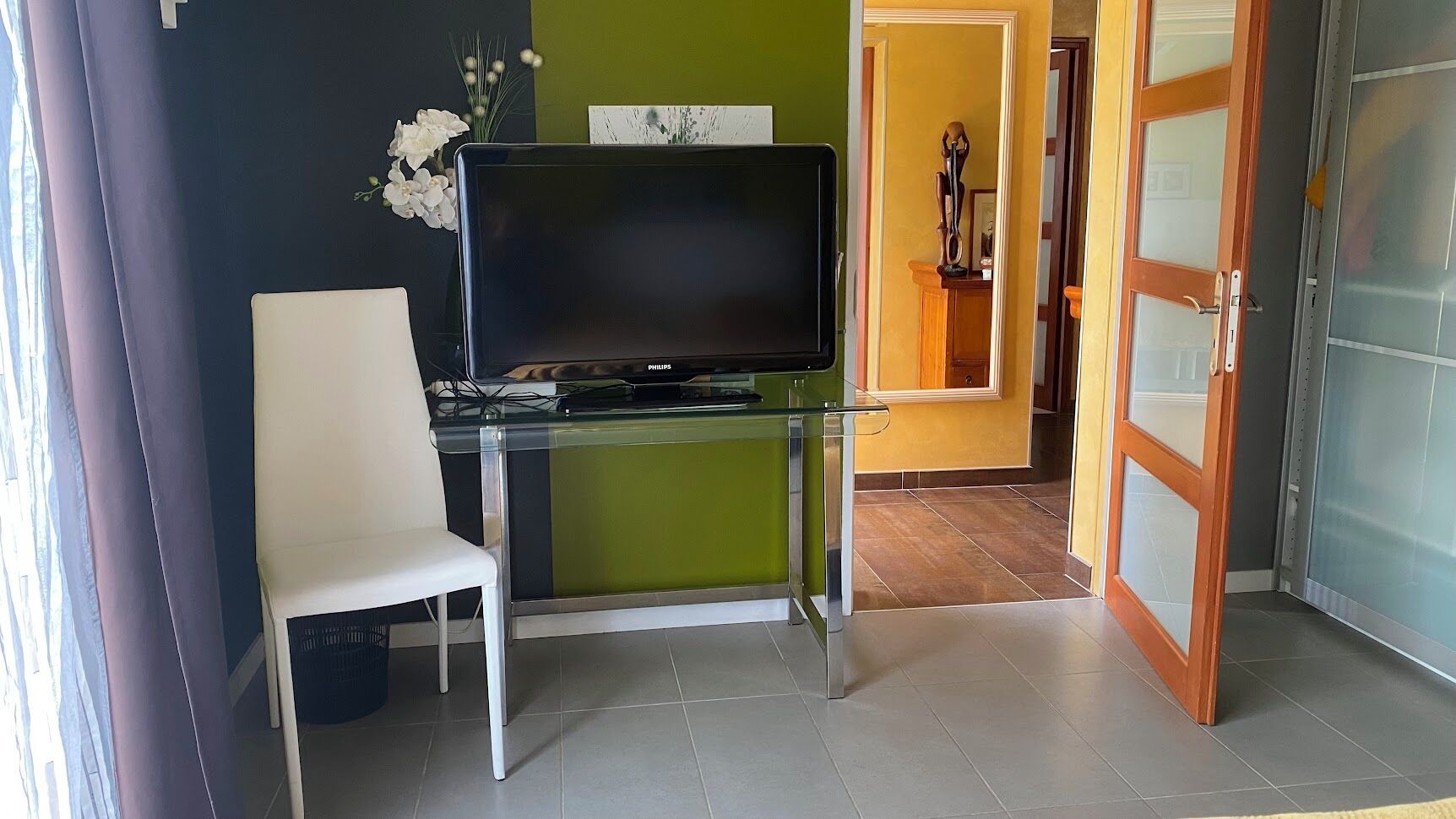 A room with a glass TV stand, a white chair, and a green wall.