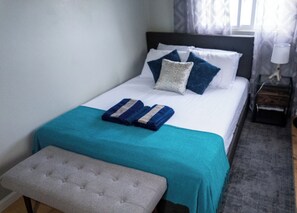 Un letto rifatto con una coperta color petrolio, due cuscini e una coperta piegata. Ai piedi del letto, una piccola panca di legno.