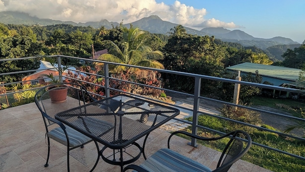 Une terrasse avec une table et des chaises en métal, donnant sur un paysage luxuriant avec des montagnes et un ciel dégagé.