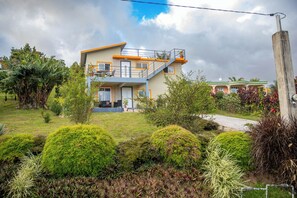 Une maison à deux étages dotée d’un balcon, entourée de jardins bien entretenus et sous un ciel dégagé.