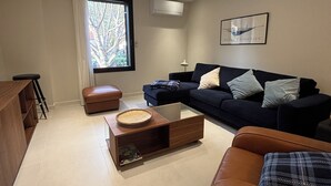 A modern living room with a dark blue sofa, a brown leather couch, and a glass coffee table. There is a window with a view of trees and a framed picture on the wall. - La Grange, in the heart of the village (Argelès-sur-Mer)
