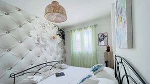 Une chambre à coucher avec une tête de lit capitonnée, un lit recouvert de draps blancs, un luminaire suspendu en rotin, un rideau vert et un tableau encadré accroché au mur.