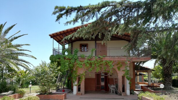 Une maison à deux étages avec un balcon, entourée d’un jardin en briques et de palmiers.