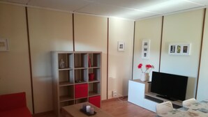 Un salon avec un plancher en bois, un canapé rouge, une table en bois, un téléviseur posé sur un meuble et une bibliothèque avec des boîtes de rangement rouges et blanches.