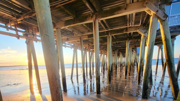 Un muelle con vigas y columnas de madera que se extiende sobre una playa arenosa.