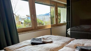 Une chambre avec deux lits, une grande fenêtre donnant sur un paysage verdoyant et une montagne, et une tête de lit de couleur sombre.
