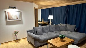 Un salon moderne avec un canapé d’angle gris, une table basse en bois et un petit pouf rond. Une lampe et un tableau encadré sont posés sur une table d’appoint.