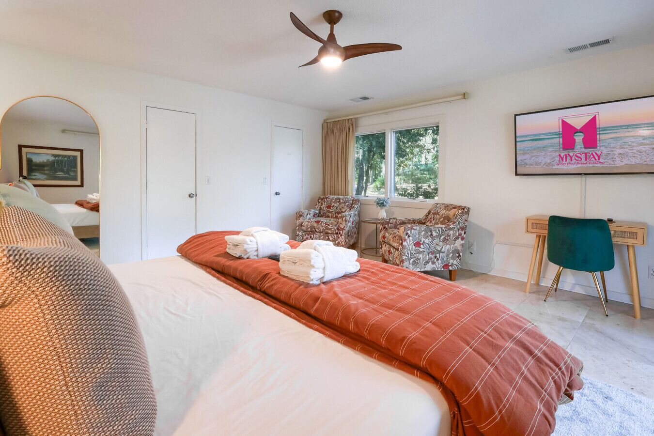 A hotel room with a large bed, a desk with a chair, a ceiling fan, and a window with curtains.
