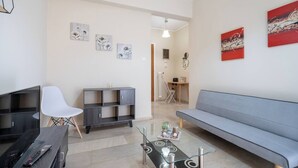 Een moderne woonkamer met een grijze bank, een witte stoel, een televisie op een houten dressoir, een glazen salontafel en een grijze boekenkast.