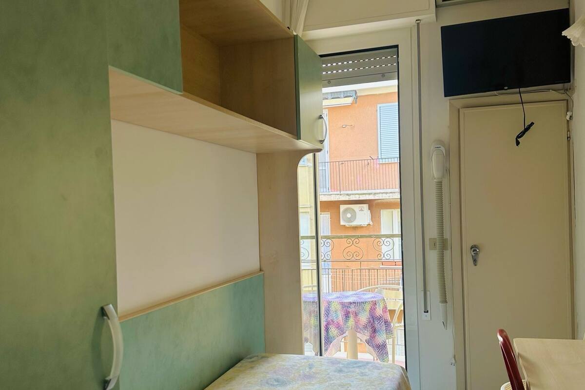 A room with a bed, a desk, and a chair. There is a window with a view of buildings outside.